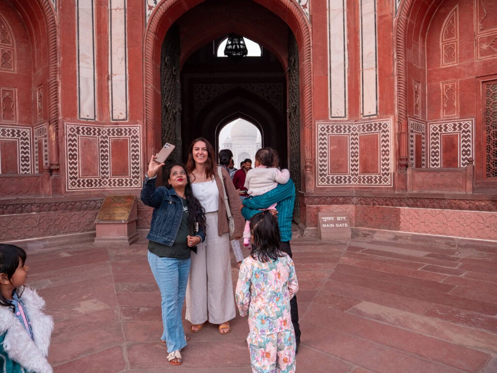 Red sandstone archway frames a view of the Taj Mahal in the distance with visitors taking selfies in front.