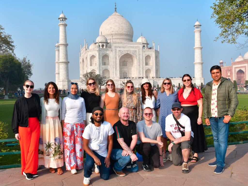Group of travellers pose together in front of the Taj Mahal with the monument clearly visible behind them. The photo captures a shared highlight of the One Life Adventures India Classic tour