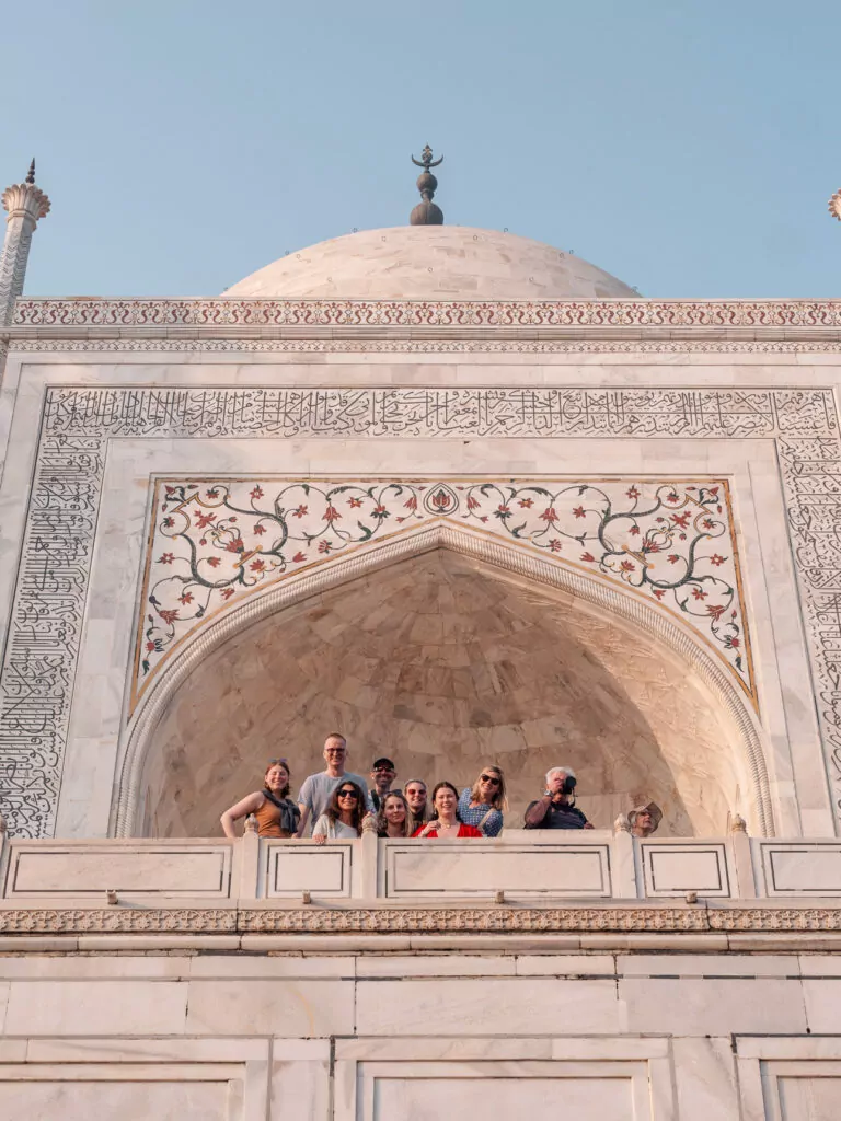 Travellers stand inside a large arched balcony with intricate marble patterns overlooking the Taj Mahal complex. The framing highlights detailed craftsmanship