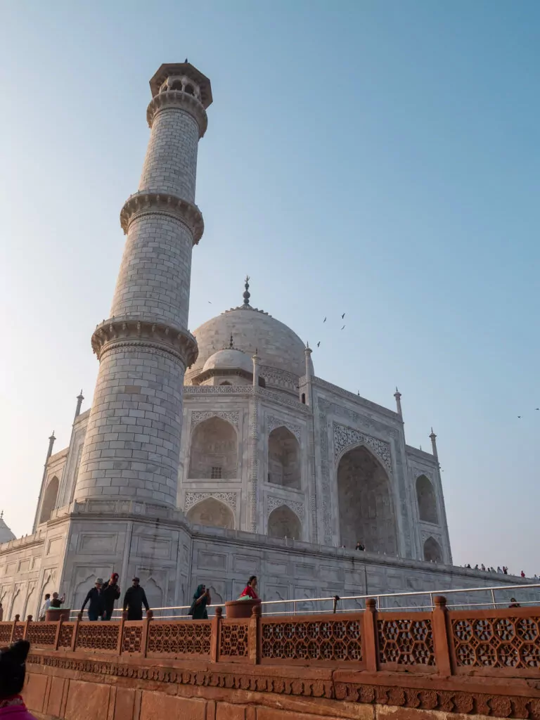 Side view of the Taj Mahal with one of its tall minarets rising beside the white marble structure. Visitors walk along the base under a clear sky.c