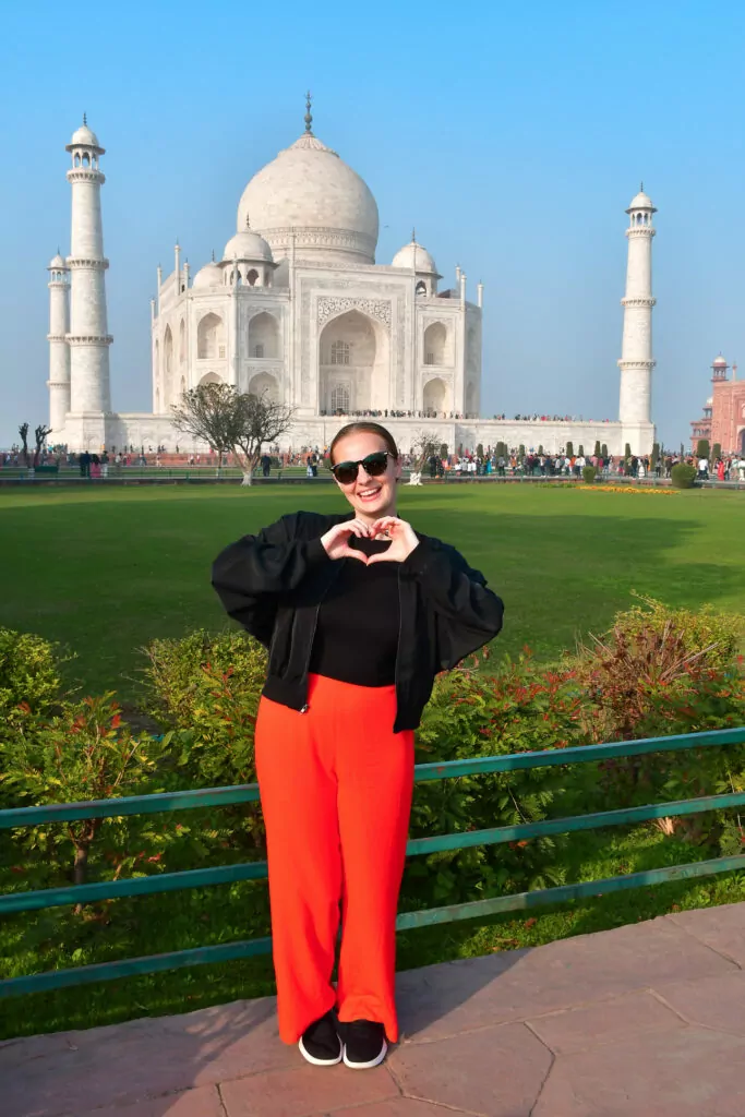 A traveller stands smiling in front of the Taj Mahal with green gardens and clear skies behind. The pose captures a memorable moment during the One Life Adventures India Classic tour
