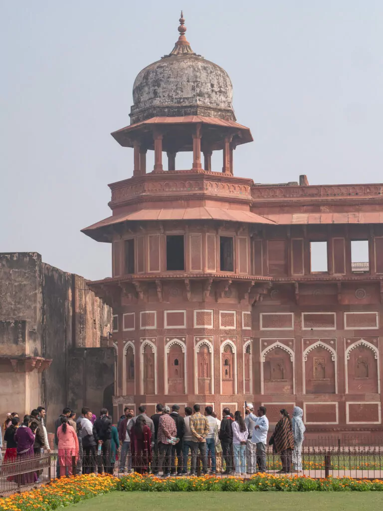 Tall red sandstone tower in Agra with layered architectural details and a crowd gathered at the base. The structure rises above the surrounding complex