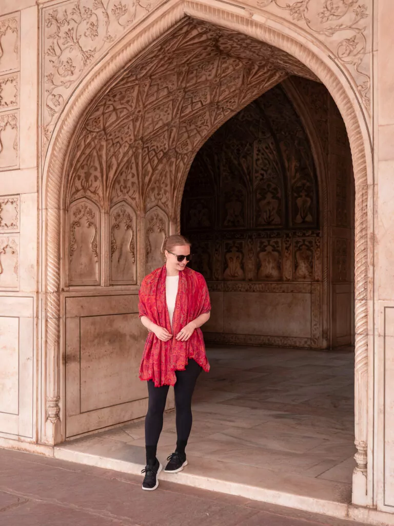 A traveller stands beneath an intricately carved stone archway wearing a red shawl. The framing highlights the detailed craftsmanship of the historic building