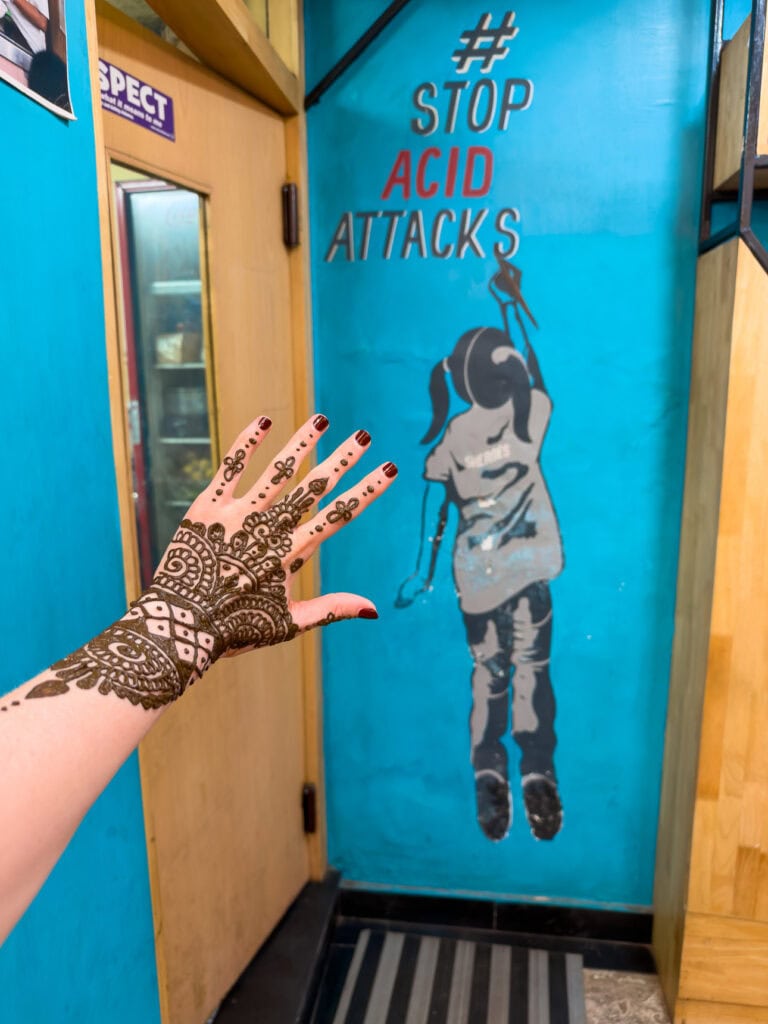 A hand with fresh henna is held up in front of a mural reading "ACID ATTACKS" at Sheroes Cafe in Agra. The powerful artwork highlights the cafe’s mission supporting survivors