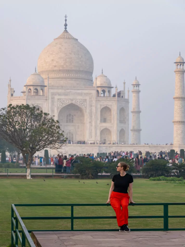 The Taj Mahal seen from a distance across a grassy area with a solo female traveller sitting on the fence in the foreground. The white marble structure stands out against a hazy sky