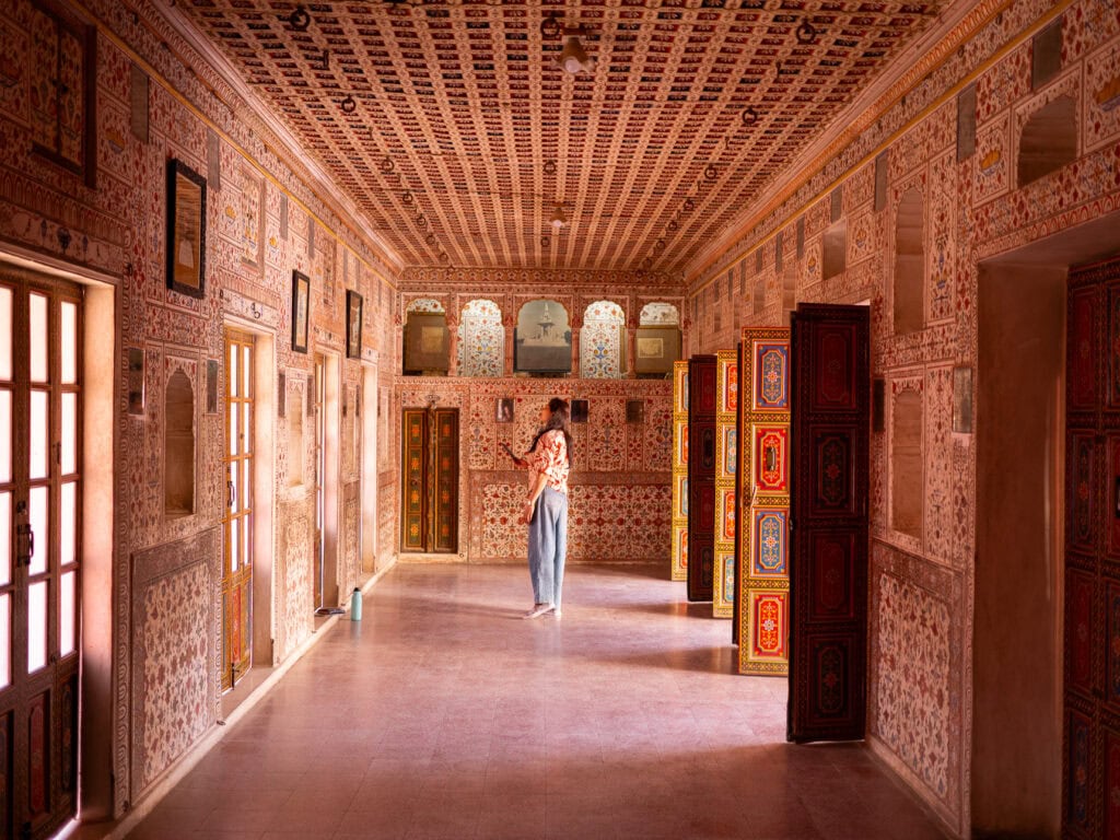 Ornate interior corridor inside a fort in Bikaner with pink sandstone walls, carved details, and arched doorways. A traveller walks through the historic space
