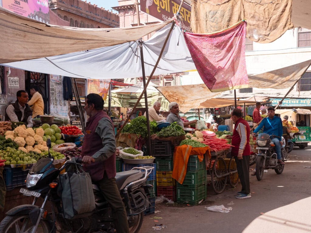 Busy street market in Bikaner with stalls covered in fabric and vendors selling goods. People walk between narrow paths lined with colourful items and local produce.