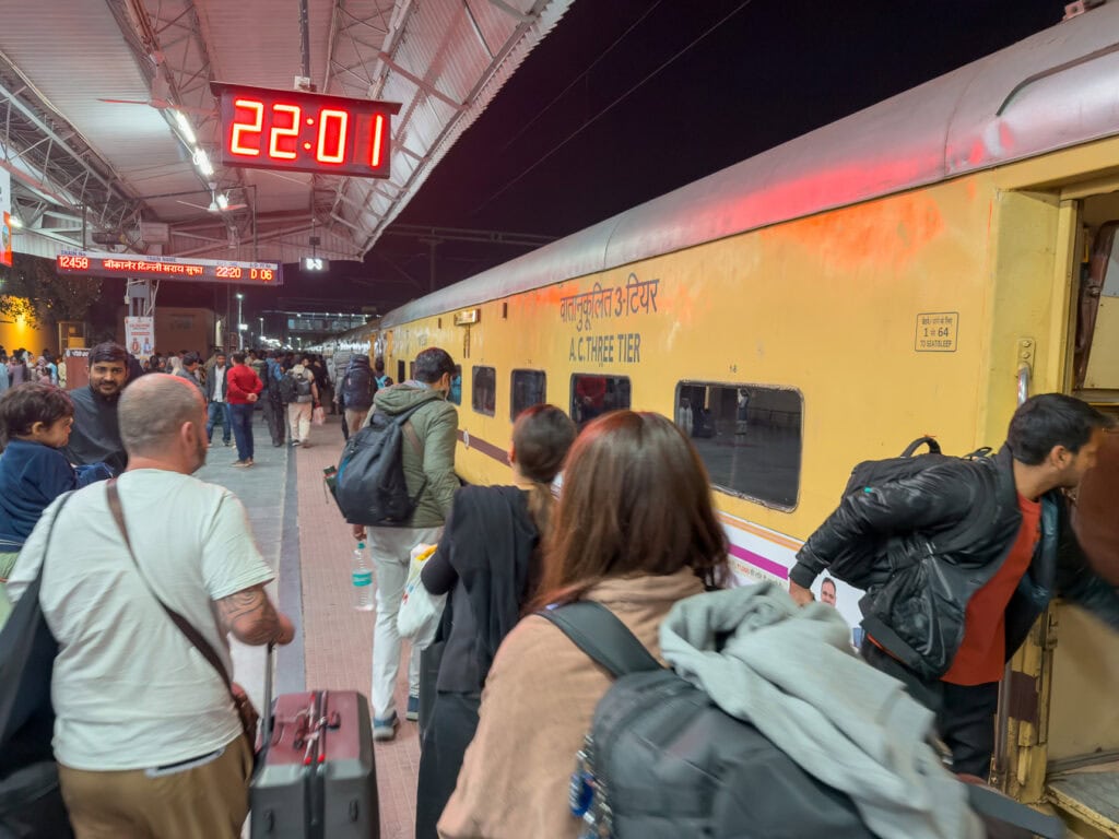 Travellers stand on a train platform next to a yellow train under station lights at night. A digital clock reads "22:01" as people prepare to board.