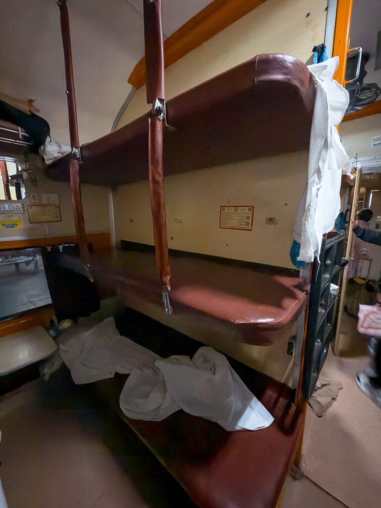 Interior of an overnight train cabin with narrow bunks and bedding stacked in tiers. Travellers rest and store their bags here during a long journey across India.