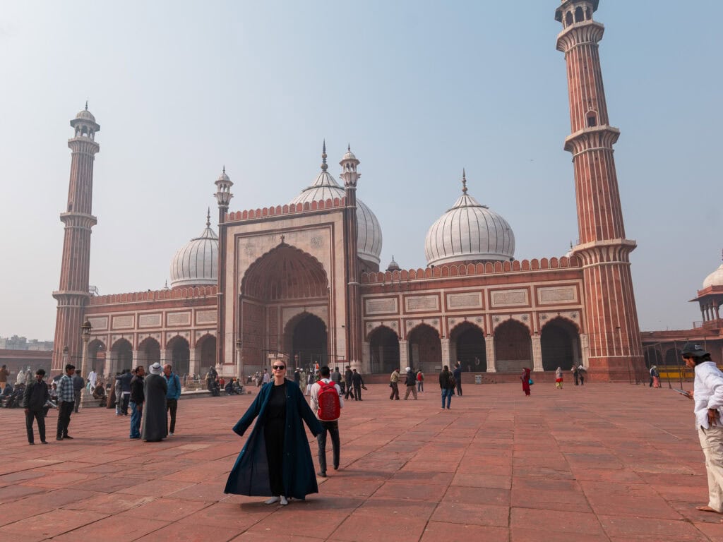 Large mosque in Delhi with domes, minarets, and a wide open courtyard filled with visitors. The historic structure stands under a clear sky