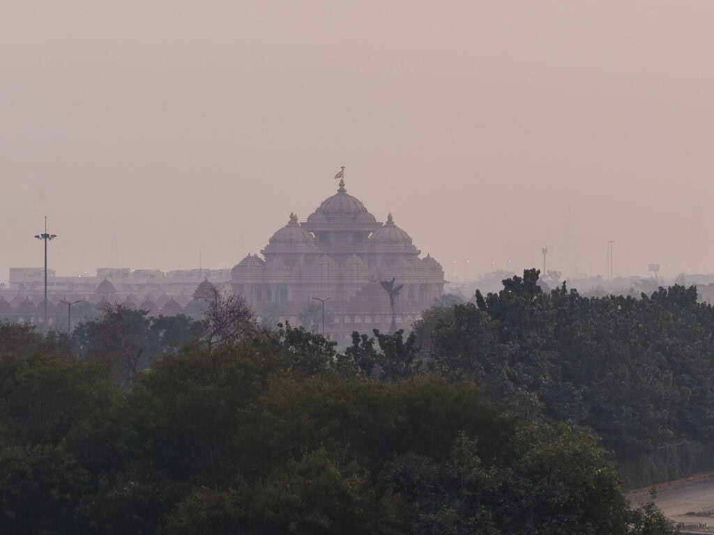 Akshardham Temple seen from a distance through light haze with its domes rising above trees. The large Hindu temple stands out against the skyline in Delhi