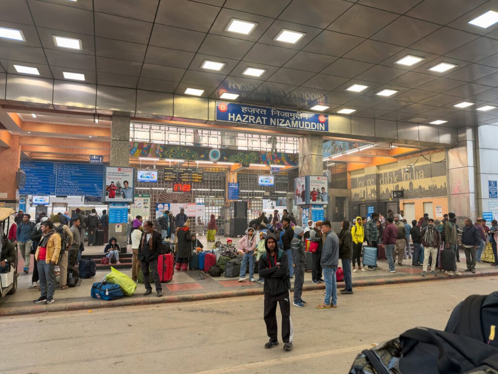 Busy train station in Delhi with travellers, luggage, and bright overhead lighting inside a large hall. Signs and platforms are visible as people move through the space