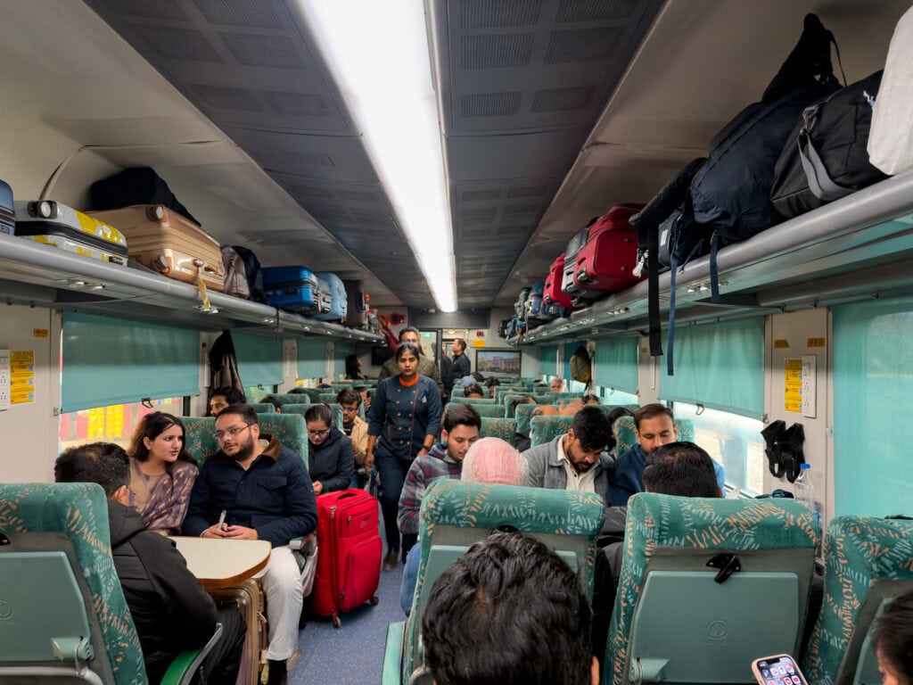 Interior of an AC third tier train carriage with bunk beds, luggage stored above, and passengers seated along the aisle. The space is busy during a journey from Delhi to Agra on the One Life Adventures India Classic tour