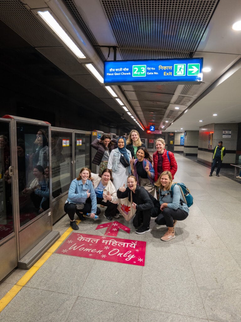 A group of travellers pose on a Delhi Metro platform with a train stopped beside them and signage overhead. The moment captures the start of an India tour with One Life Adventures
