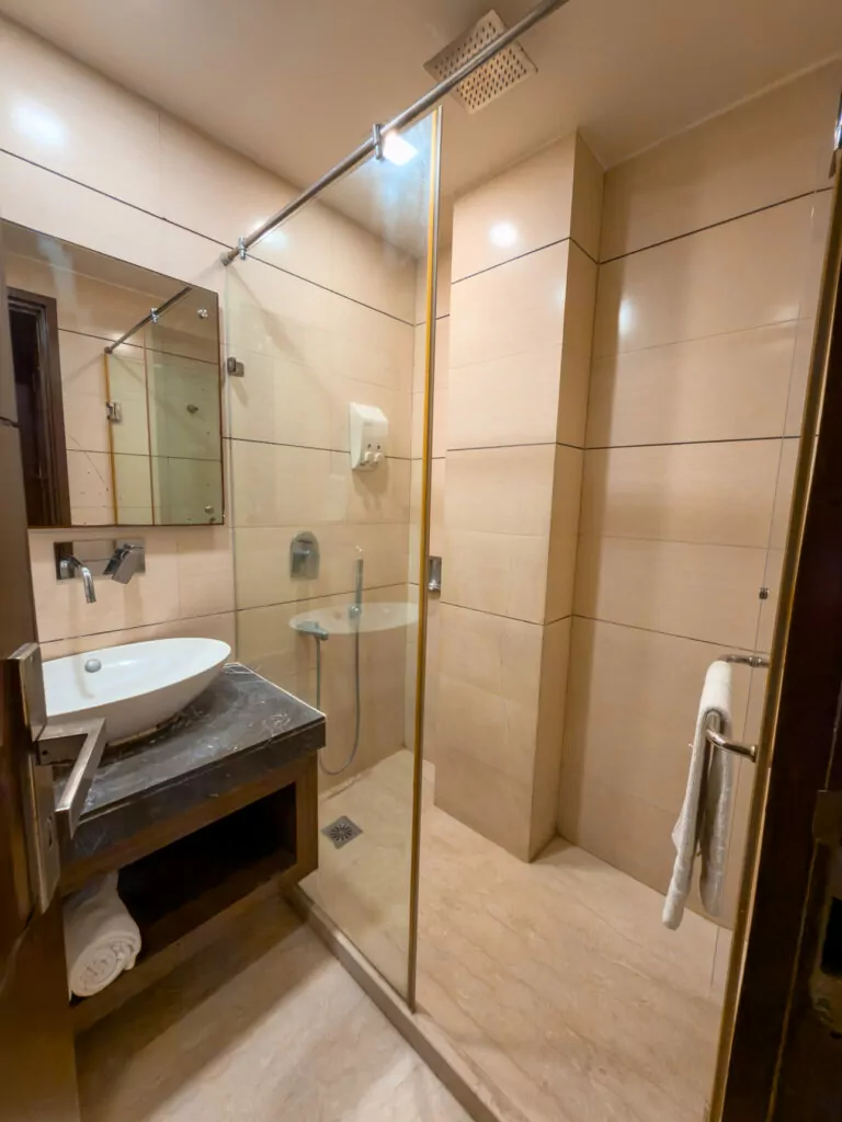 Bathroom at Taj Princess hotel in Delhi with a glass shower, sink, and tiled walls