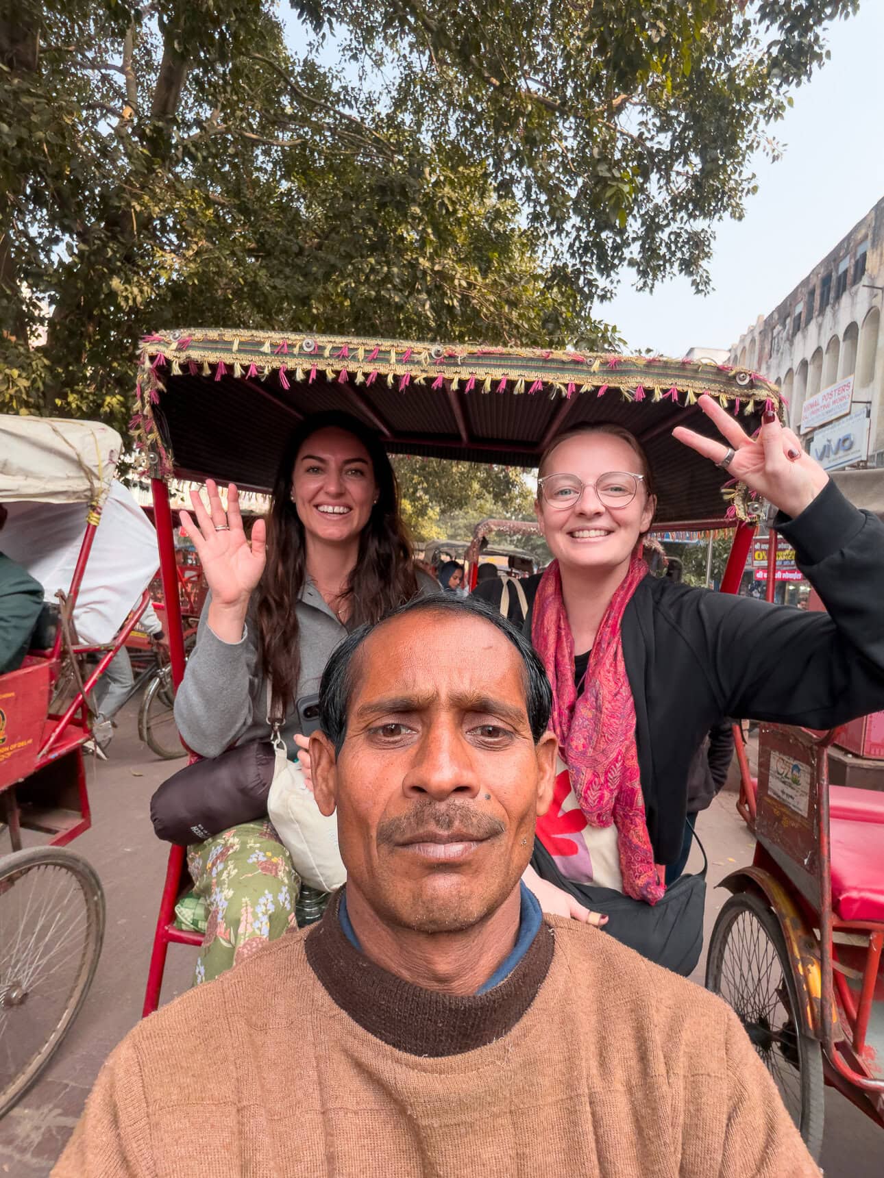 A rickshaw driver takes a selfie with two travellers sitting in the back seat smiling and waving. The candid moment captures a fun part of travelling through Delhi