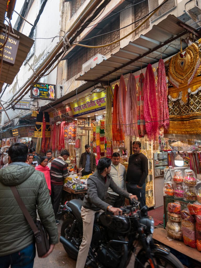Busy street in Jaipur lined with colourful shops, hanging textiles, and people walking between motorbikes