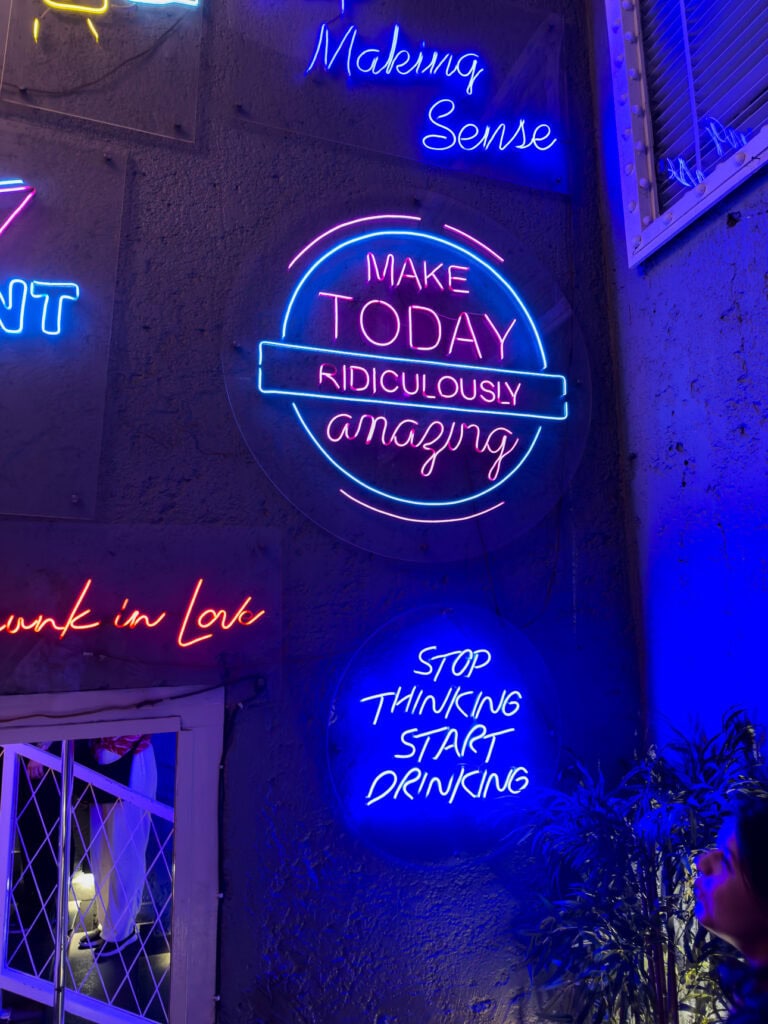 Bright neon signs glow in blue and pink inside a bar in Delhi with phrases like "Make today ridiculously amazing" and "Stop thinking start drinking." The vibrant nightlife scene is part of the One Life Adventures India experience.