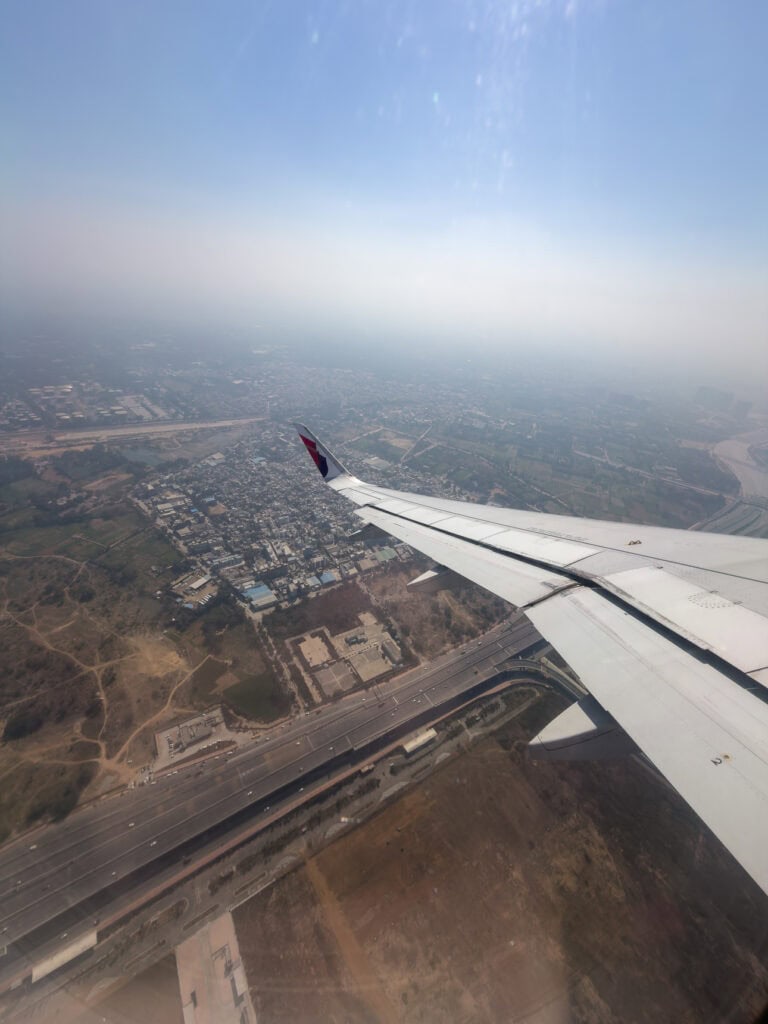 View from an airplane window flying over Delhi with buildings and roads fading into haze below. The wing stretches across the frame during arrival or departure in India