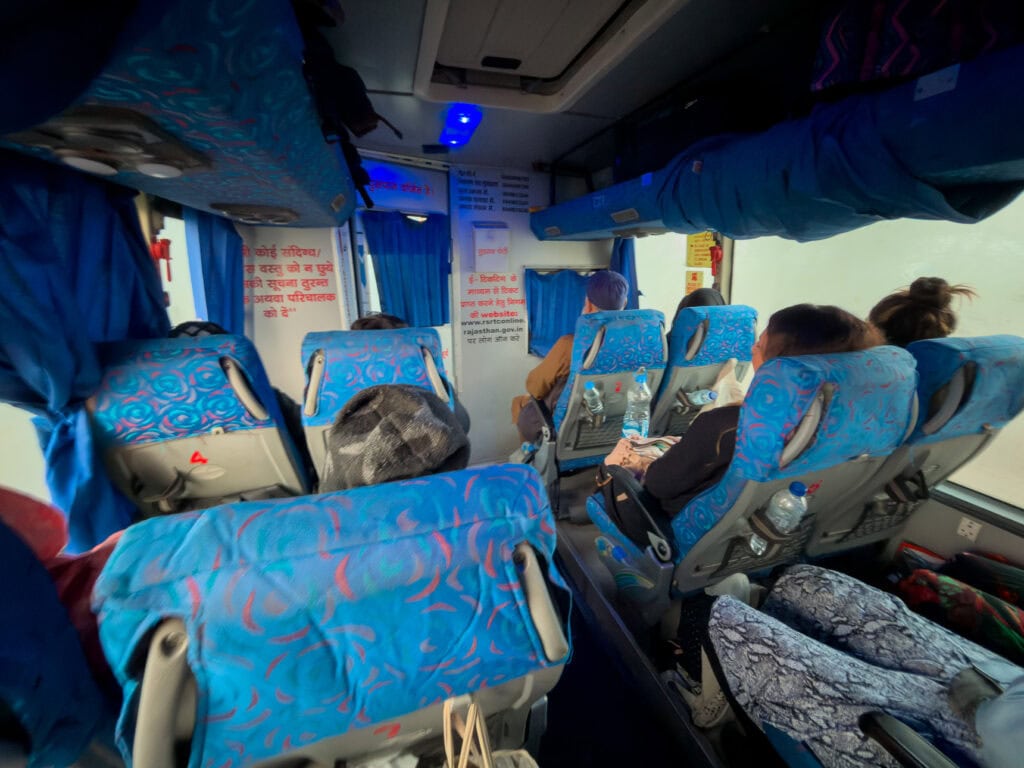 Rows of blue seats inside a coach bus with travellers seated and resting during transit. The journey forms part of a group India tour itinerary
