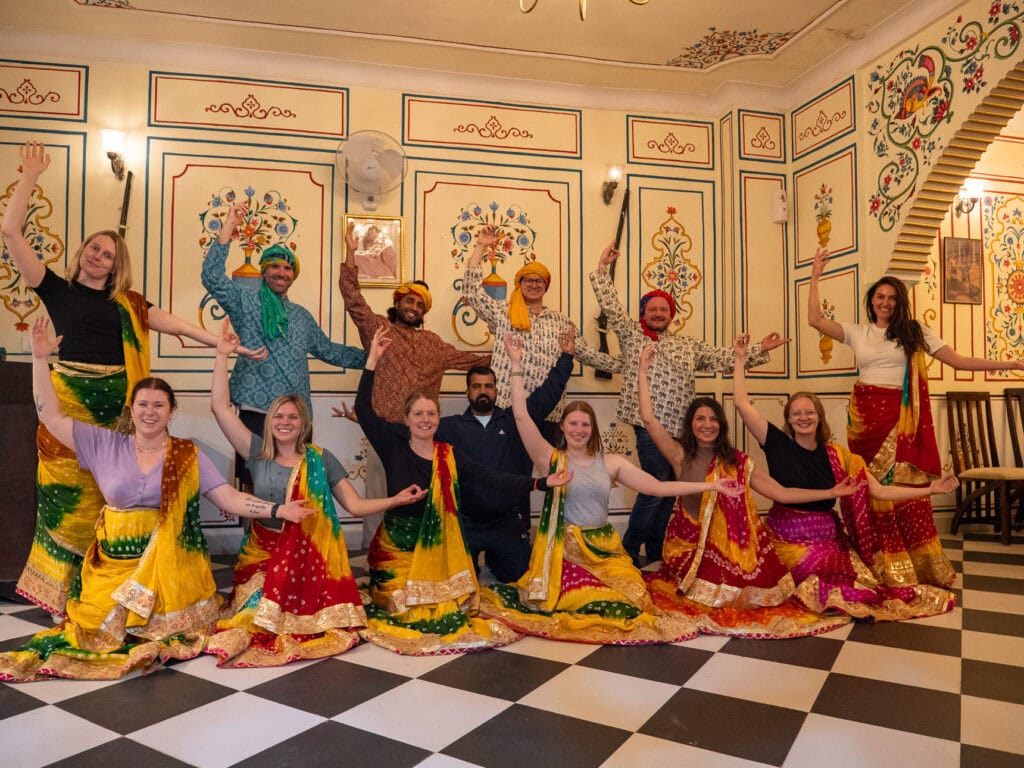A group of travellers pose with local performers in colourful traditional outfits inside a decorated hall. The lively scene captures a cultural experience during the One Life Adventures India tour