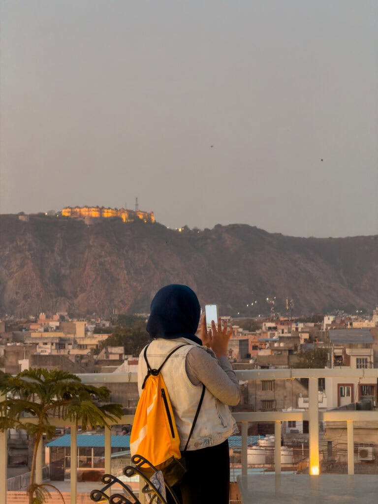 A traveller looks out over Jaipur from a rooftop with a hilltop fort visible in the distance under soft evening light