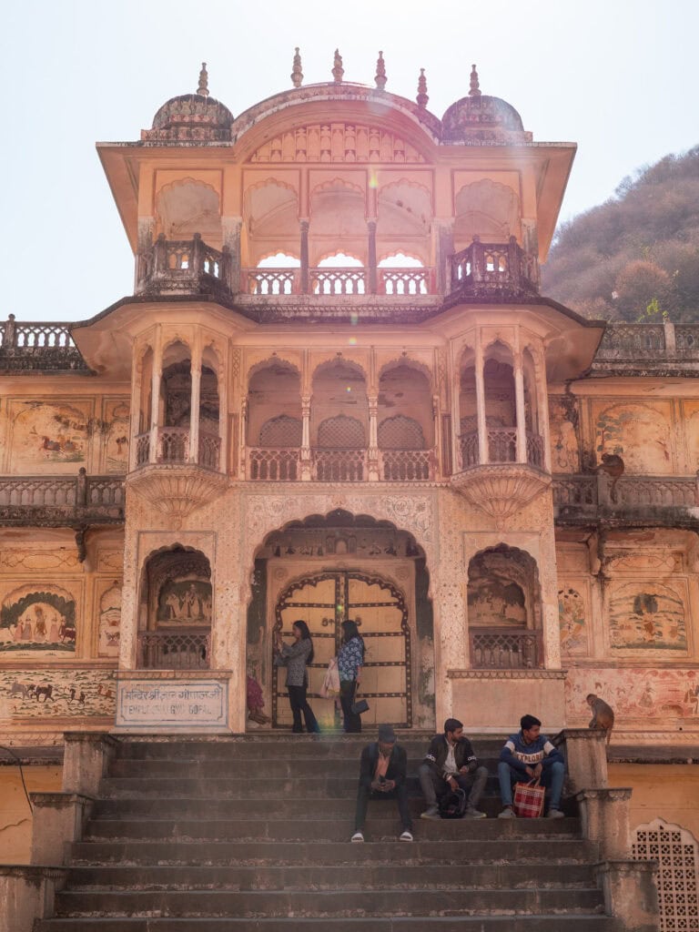 People walk through Galta Ji Monkey Temple with historic pink sandstone structures and steps leading through the complex. The setting is surrounded by hills and traditional architecture