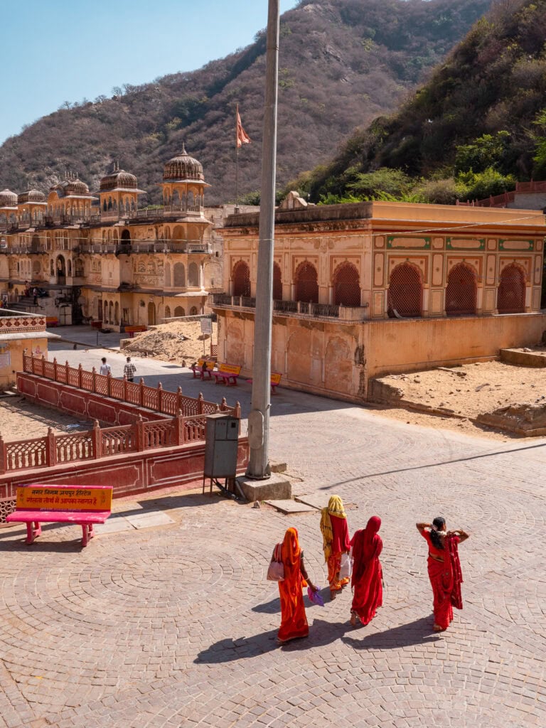 View over Galta Ji Monkey Temple with historic buildings, courtyards, and hills surrounding the complex. A few people in bright clothing walk through the open space below