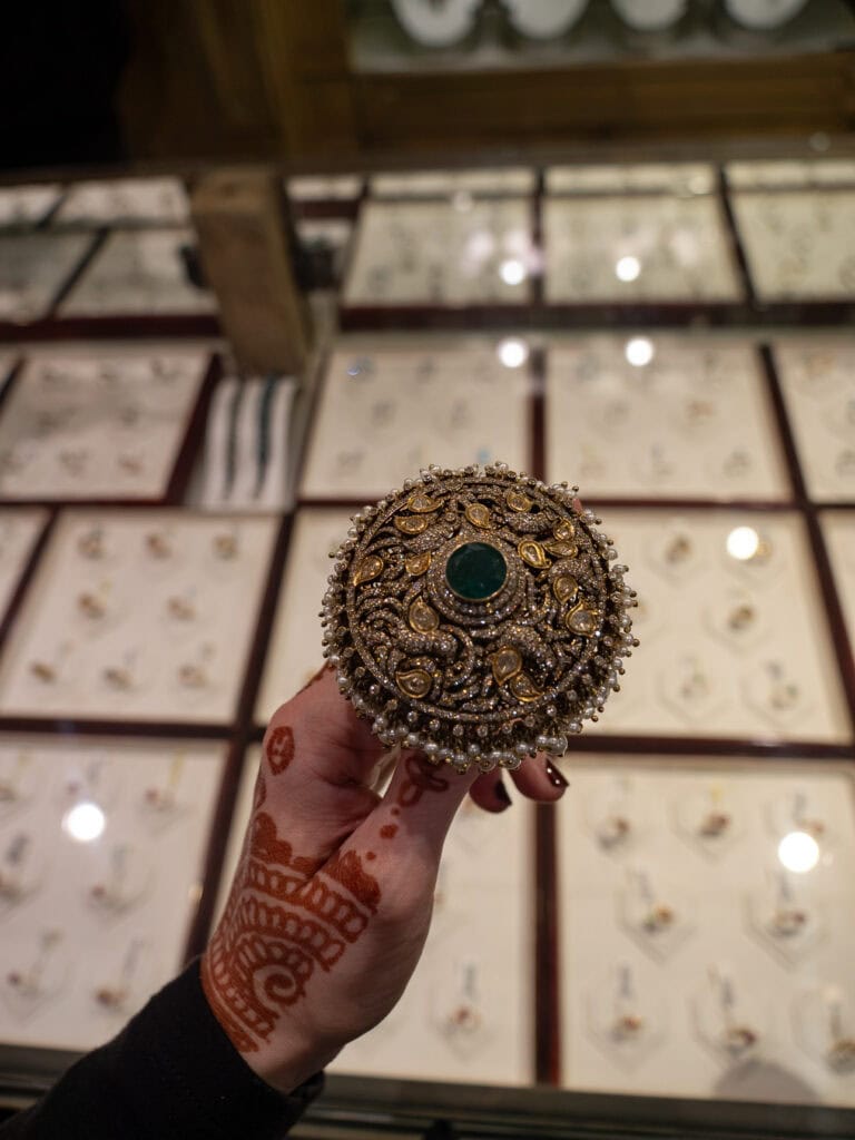 A hand holds up an ornate ring with a large green stone and intricate gold detailing inside a jewellery shop in Jaipur. The craftsmanship reflects traditional Indian design.