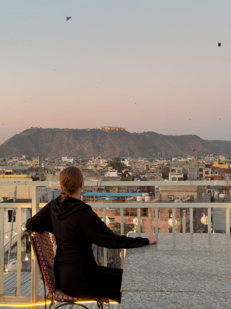 A traveller looks out over Jaipur from a rooftop with buildings and hills stretching into the distance at sunset. The soft light creates a calm end to the day on an India tour.