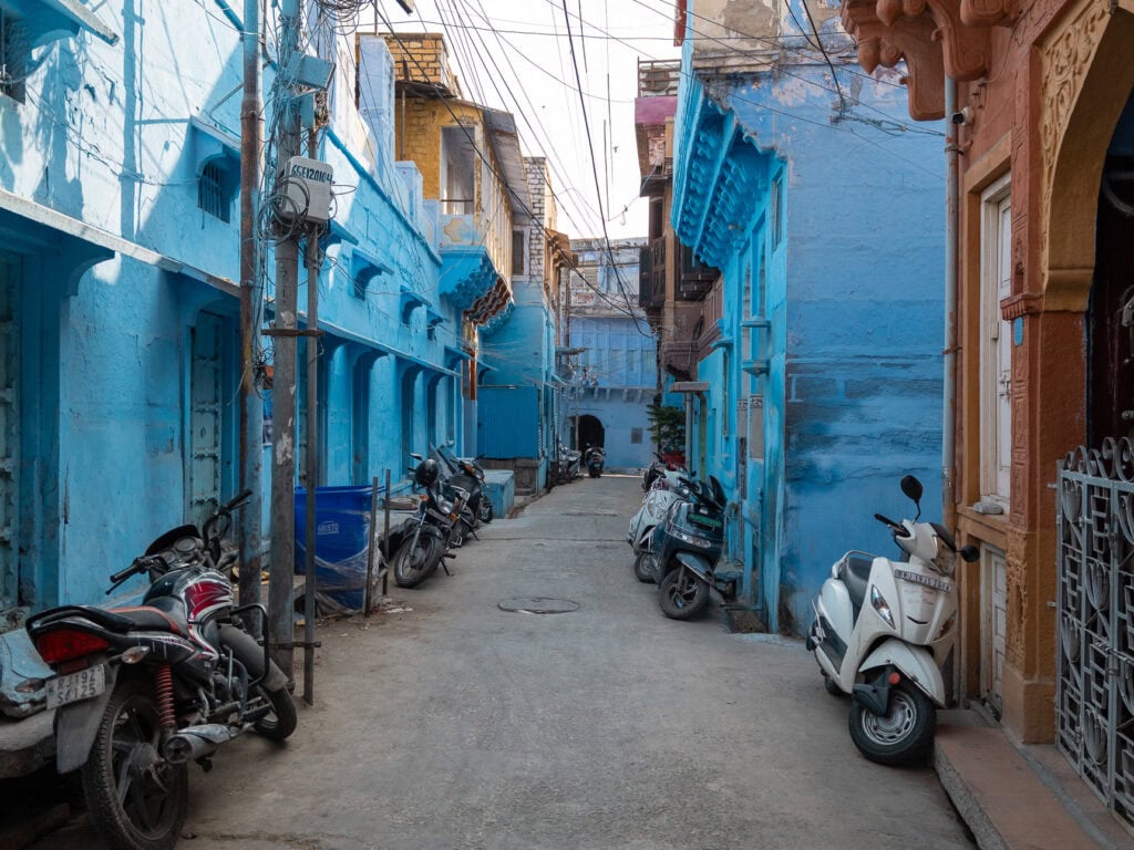 Narrow alley in Jodhpur lined with bright blue buildings, doorways, and parked motorbikes. The colour gives the city its distinctive character