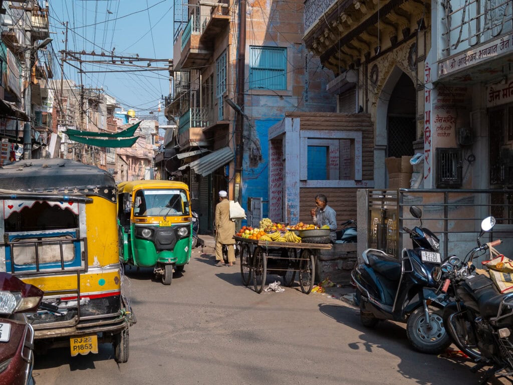 Everyday street life in Jodhpur with auto rickshaws, pedestrians, and small shops along the road.