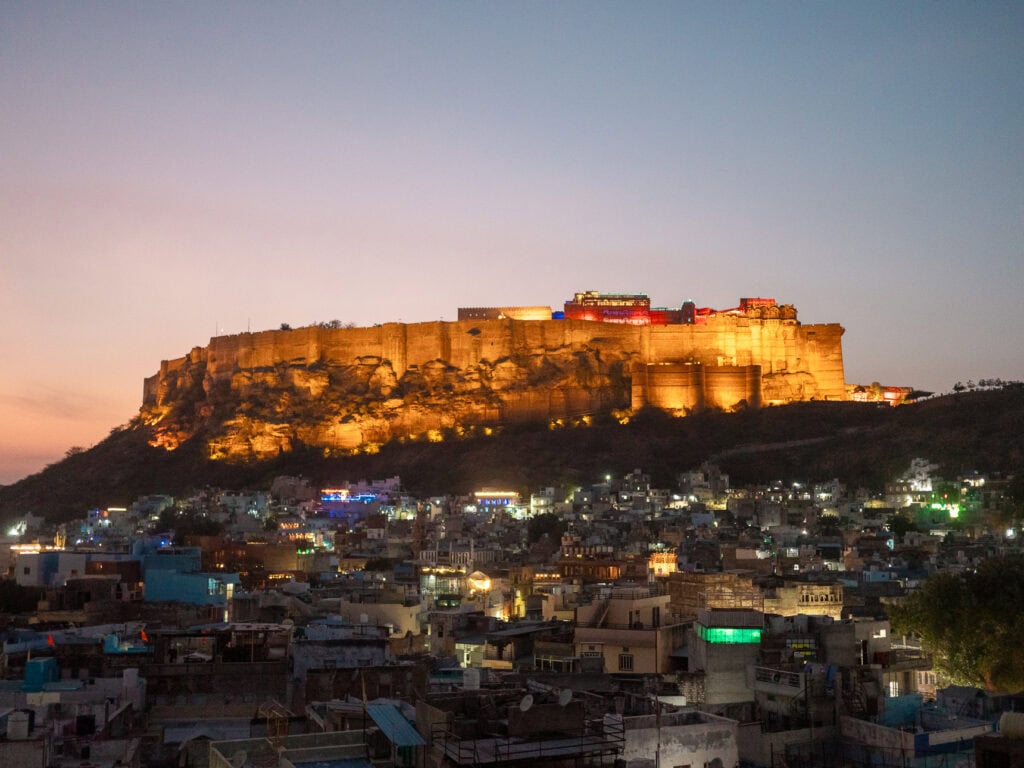 Mehrangarh Fort illuminated at night with warm golden lights glowing against the dark sky. The city of Jodhpur spreads out below in the distance