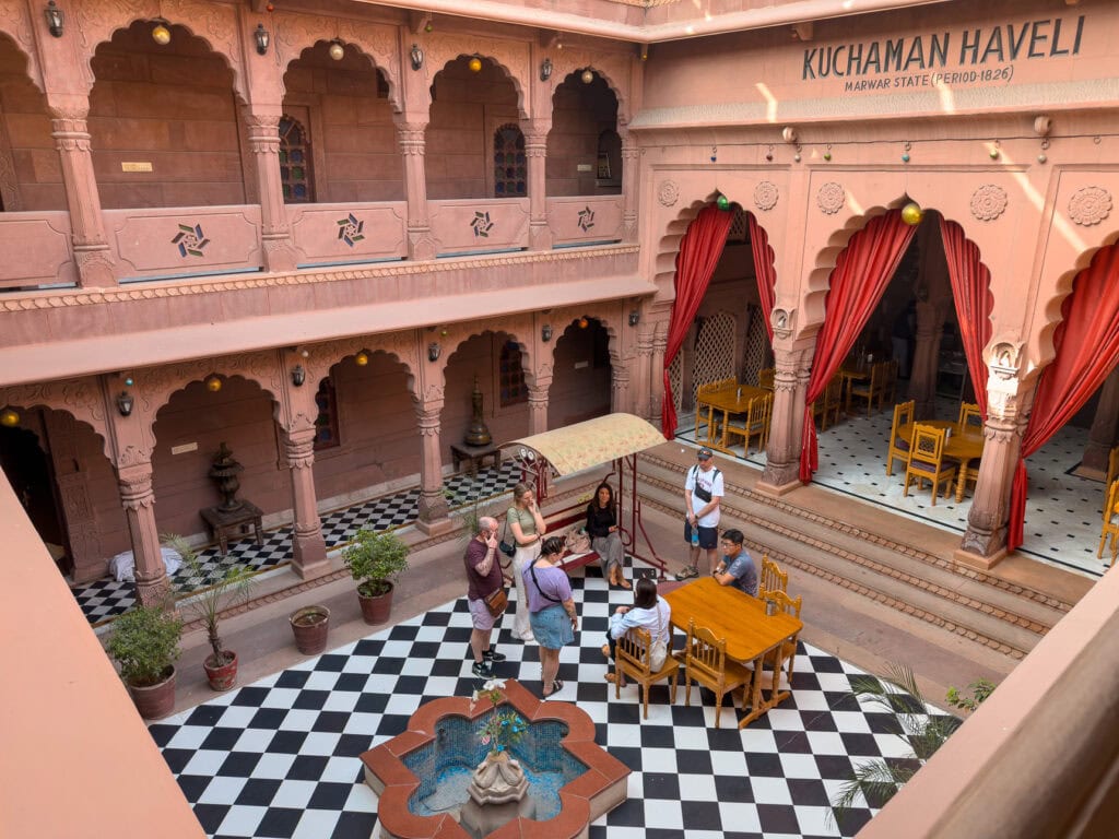 Courtyard at Kuchaman Haveli in Jodhpur with arched balconies, red curtains, and a patterned tiled floor. Guests gather around tables in the centre of the space