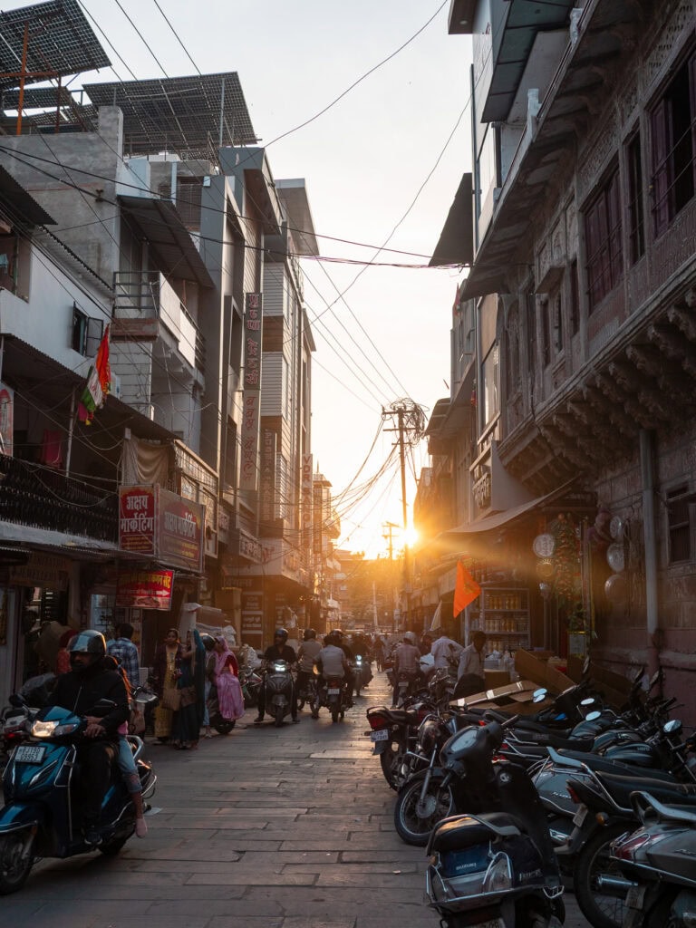 Narrow street in Jodhpur at sunset with warm light glowing between buildings and people walking. Motorbikes and shops line the busy lane