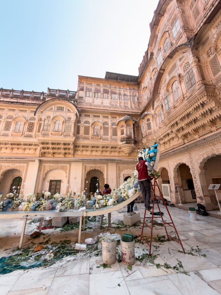 Workers arrange colourful flowers and decorations inside a fort courtyard in preparation for a wedding. The scene captures traditional celebrations within historic Indian architecture