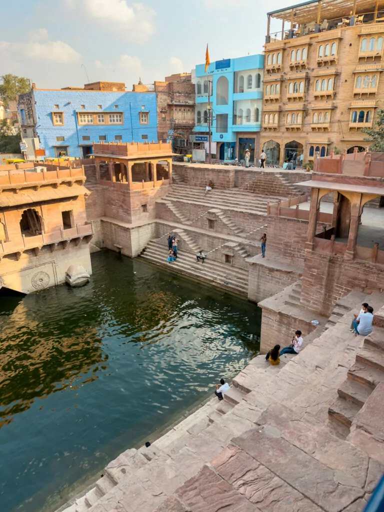 Stone stepwell in Jodhpur with geometric stairs leading down to green water. Historic buildings surround the structure creating a peaceful scene