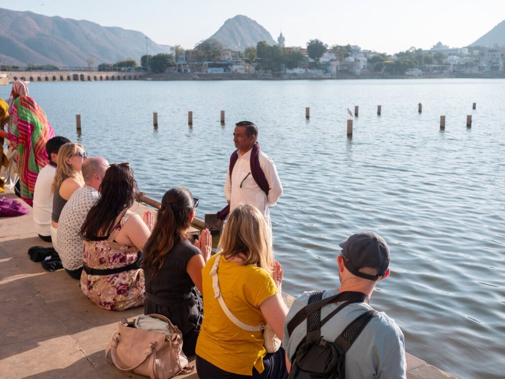 A guide speaks to a group of travellers sitting by the lake in Pushkar with hills visible in the distance. The group listens during a cultural stop on an India tour for solo travellers