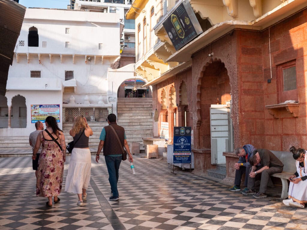 Travellers walk through a tiled courtyard in Pushkar with an arched gateway leading towards steps in the distance. The area is quiet with soft natural light