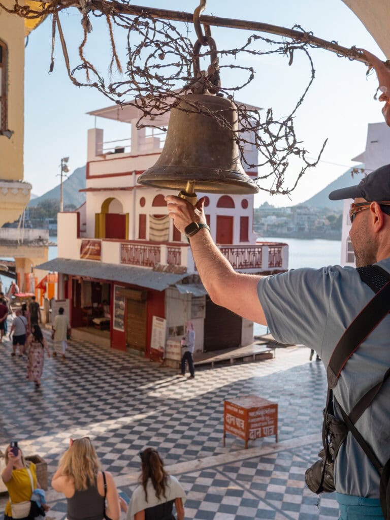 A traveller rings a large hanging bell by the lakefront in Pushkar with calm water and buildings in the background. The moment captures a peaceful cultural experience during an India tour