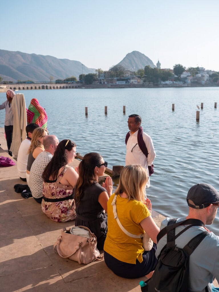 A group of travellers sit by the lake in Pushkar listening to a guide with water and hills in the background. The moment captures a peaceful cultural stop on an India small group tour itinerary.