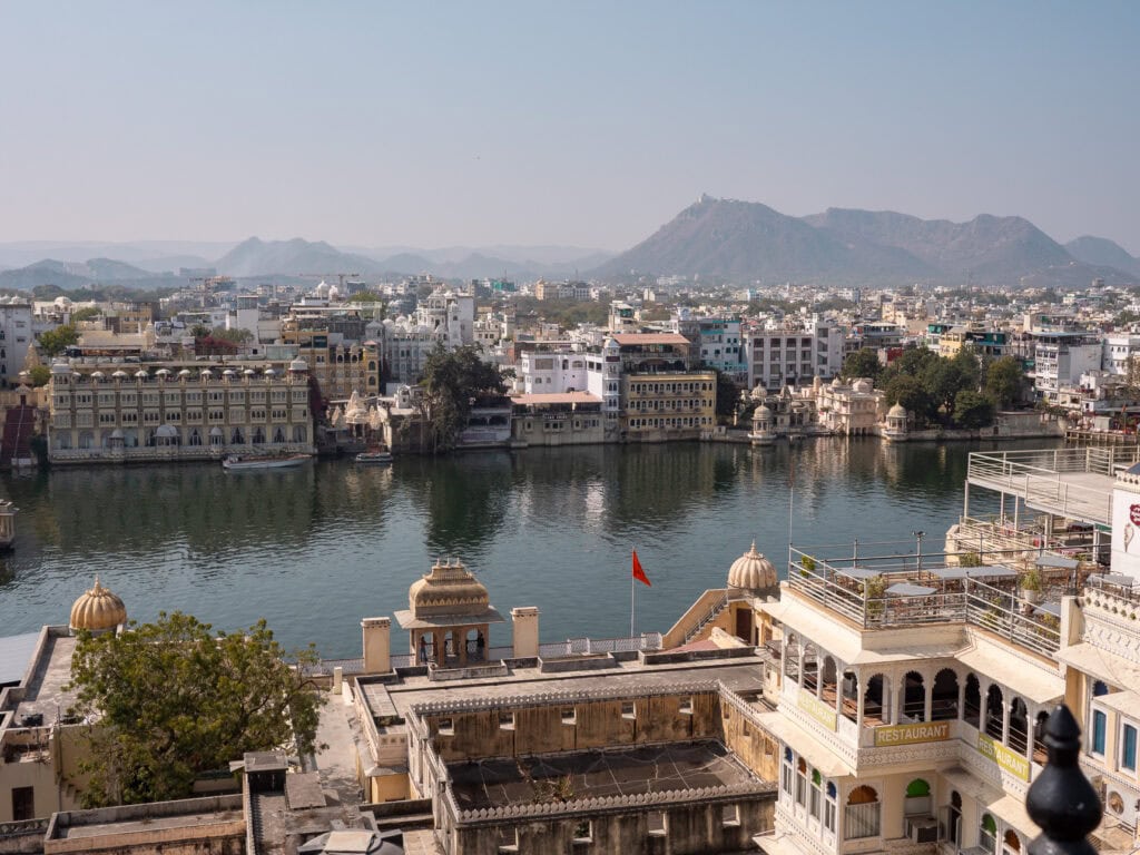 Rooftop view over Udaipur with lakes, buildings, and distant hills under a hazy sky. The elevated perspective shows the layout of the city