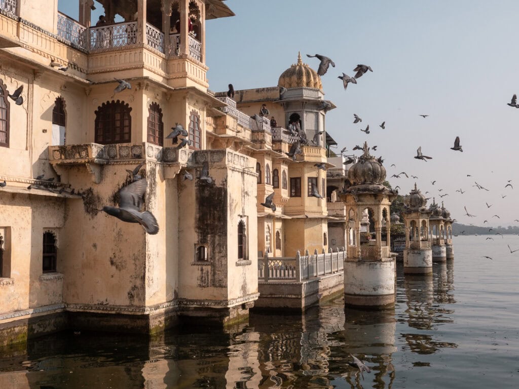 Lal Ghat in Udaipur with historic buildings lining the water’s edge and reflections in the lake. Birds fly above the calm scene at golden hour