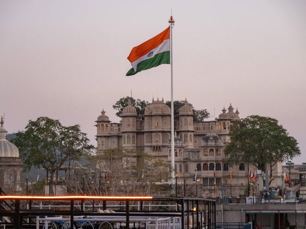 Rooftop view over Udaipur with historic buildings and a lake stretching into the distance. The Indian flag waves above the city skyline under a hazy sky