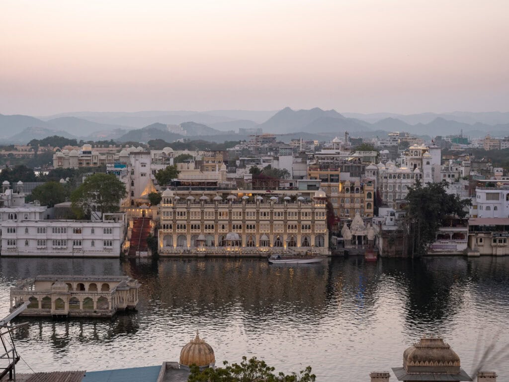 Rooftop view over Udaipur with a lake, historic buildings, and hills fading into the distance at sunset. The calm setting contrasts with the busy streets below