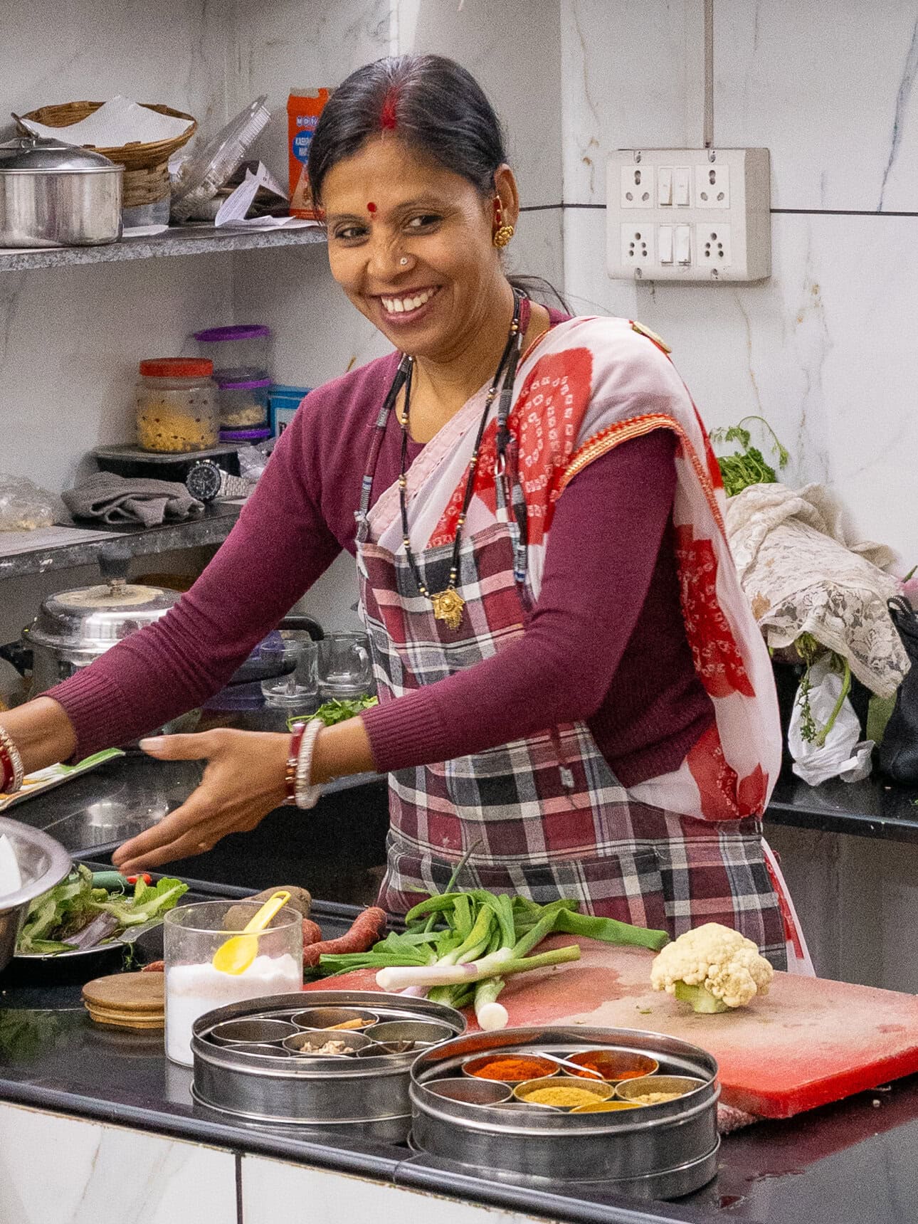 A local instructor cooks at a stove during a Udaipur cooking class guiding the process fr tourists who are watching. Ingredients and utensils are arranged nearby