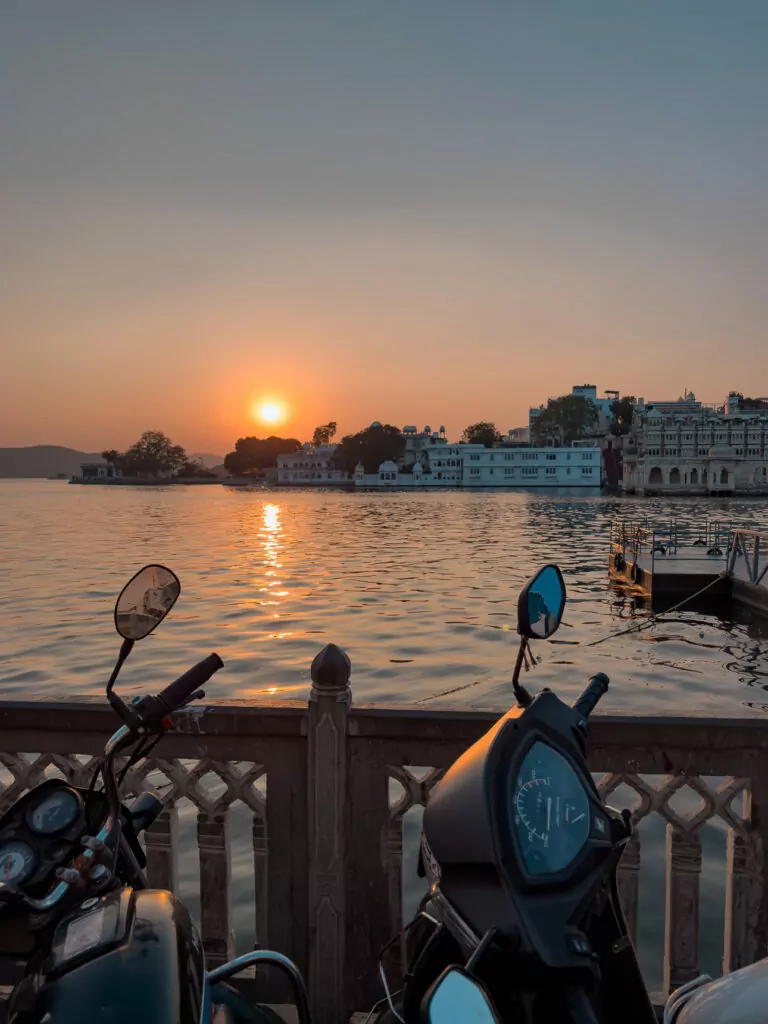 Sunset over Lake Pichola with boats and buildings silhouetted against the orange sky. The peaceful view is a highlight of Udaipur
