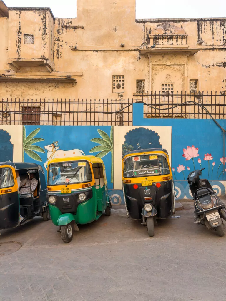 Street scene in Udaipur with colourful wall art and several tuk tuks parked along the road. The setting shows everyday life and transport in the city