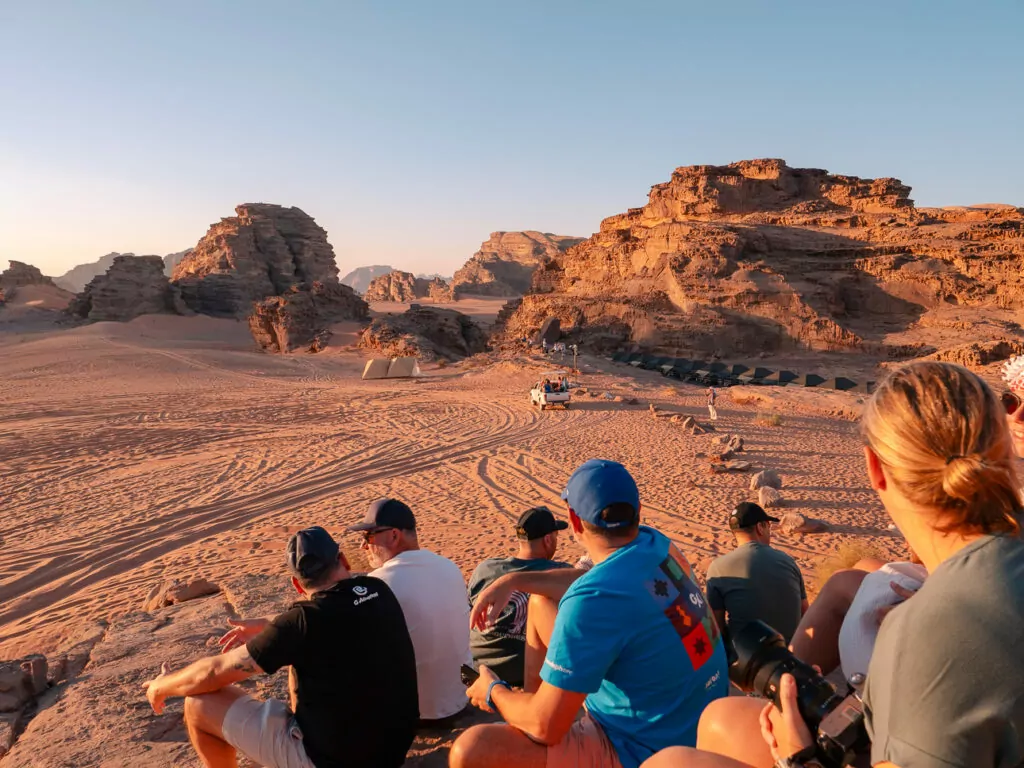 G Adventures tour group sitting together on the sand watching sunset in Wadi Rum, with desert cliffs glowing in golden light