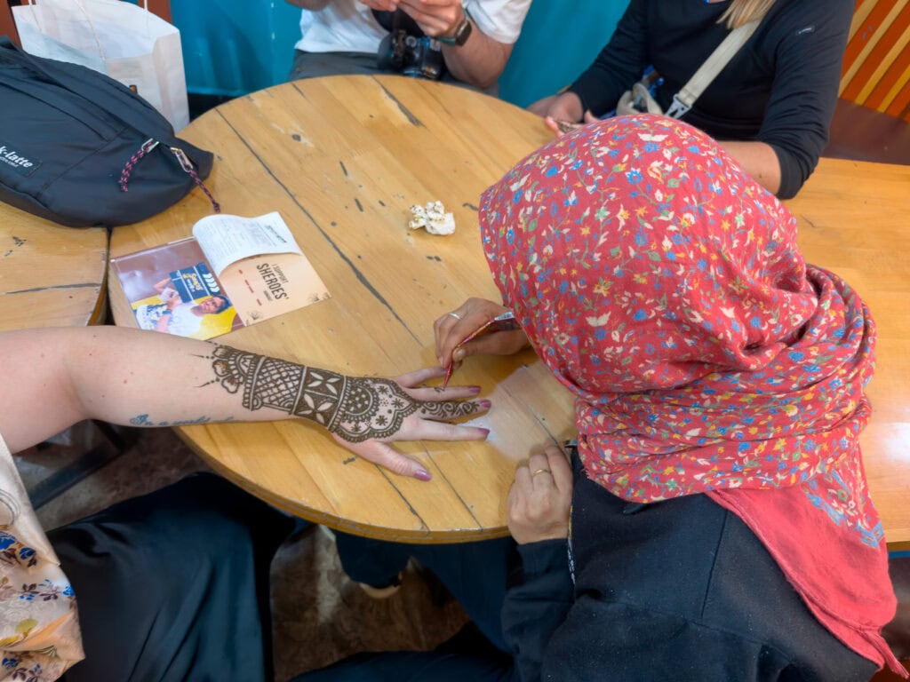 Intricate henna design being applied to a woman’s hand at a round wooden table, capturing a hands-on cultural moment